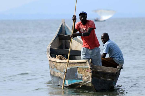 Fishing boat on water with one man sitting on the right edge and the another man, in red t-shirt, is standing and holding a long stick for navigation